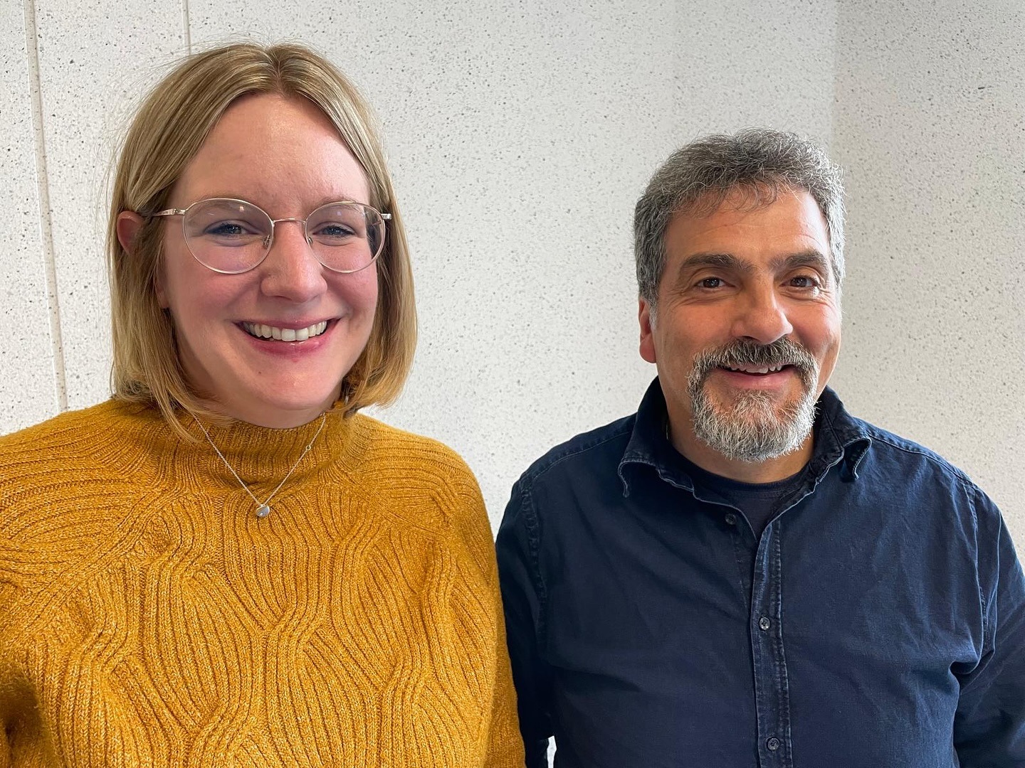 Zwei Personen stehen nebeneinander vor einer hellen Wand, eine trägt einen senfgelben Pullover, die andere ein dunkelblaues Hemd.