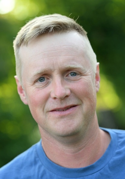 Mann mit kurzen blonden Haaren trägt ein blaues T-Shirt, unscharfer grüner Hintergrund