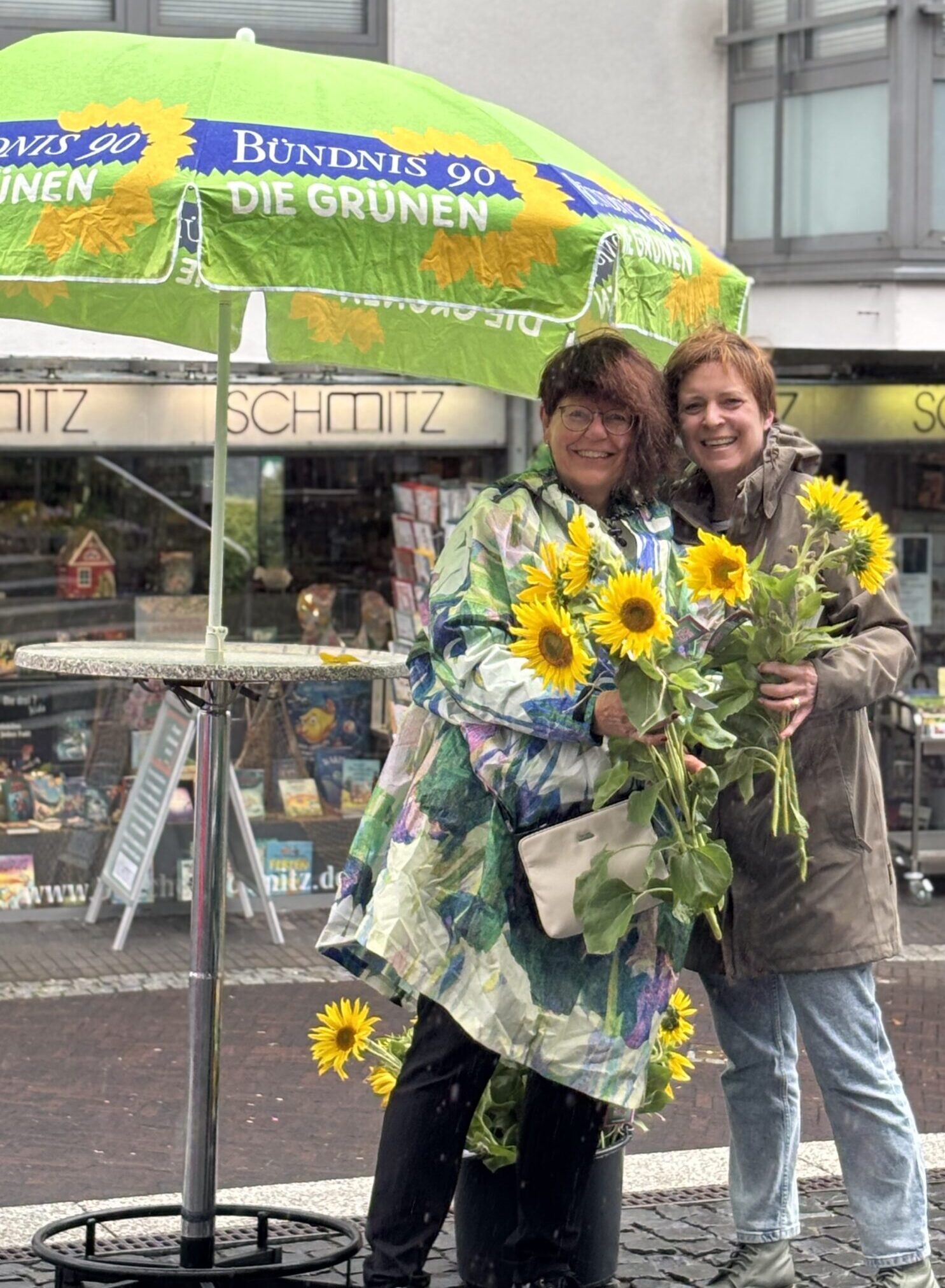 Zwei Personen stehen unter einem grünen Sonnenschirm mit Aufschrift 'Bündnis 90 Die Grünen' und halten Sonnenblumen in der Hand, im Hintergrund ein Geschäft mit dem Namen 'Schmitz'