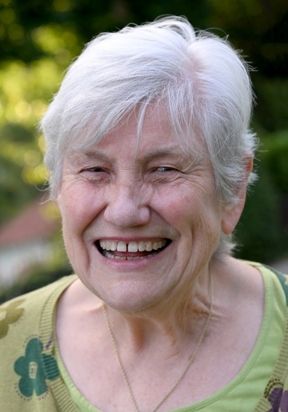 Ältere Person mit kurzen weißen Haaren, trägt grünes Oberteil mit Blumenmuster und goldene Halskette, vor grünem Hintergrund