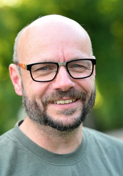 Mann mit Glatze und kurzem Bart trägt ein graues T-Shirt, unscharfer grüner Hintergrund