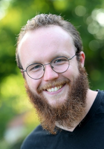 Mann mit dichtem, langem braunem Bart und lockigem Haar, trägt schwarzes T-Shirt, unscharfer grüner Hintergrund